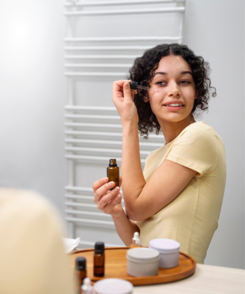 A woman with curly dark hair wearing a pale yellow t-shirt is shown in a bathroom setting, holding a small amber dropper bottle and looking into a mirror. A wooden tray with skincare jars sits on the counter in the foreground.