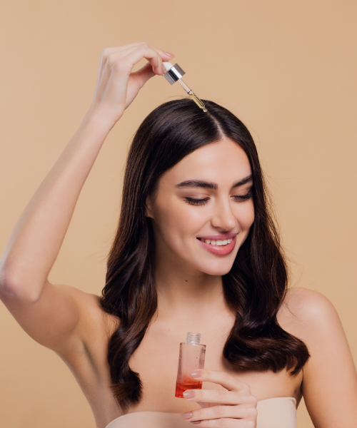 A woman with long dark brown hair applies a golden oil from a dropper to her hair, holding a small clear bottle with red liquid in her other hand, set against a solid beige background.