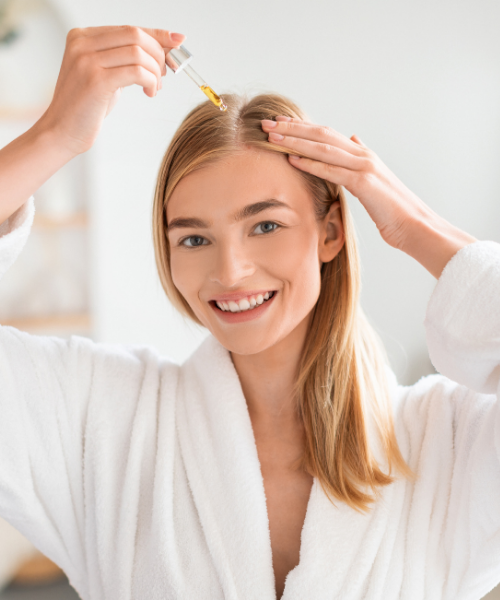 A smiling woman with blonde hair wearing a white bathrobe applies a golden-yellow hair oil from a glass dropper to the center of her hairline in a bright, clean bathroom setting.