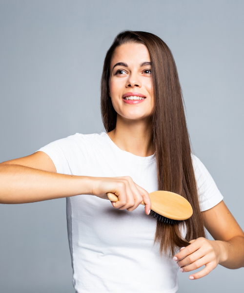 A woman with long, straight brown hair brushes her hair with a wooden hairbrush while wearing a white t-shirt against a solid gray background.