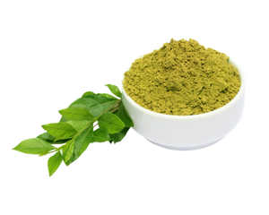A white bowl filled with green powder, shown next to fresh green leaves on a plain white background.