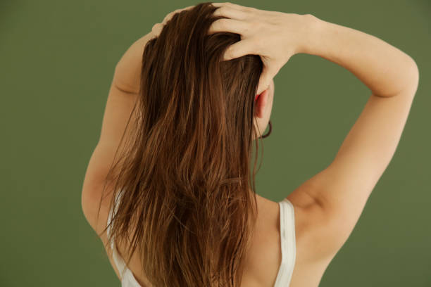 A woman with long, dark brown hair seen from behind, holding her hair in her hands against a solid olive green background.